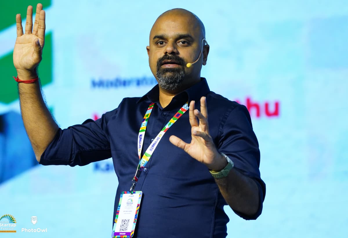 Maran speaking on stage with a headset microphone, both hands raised mid-point
