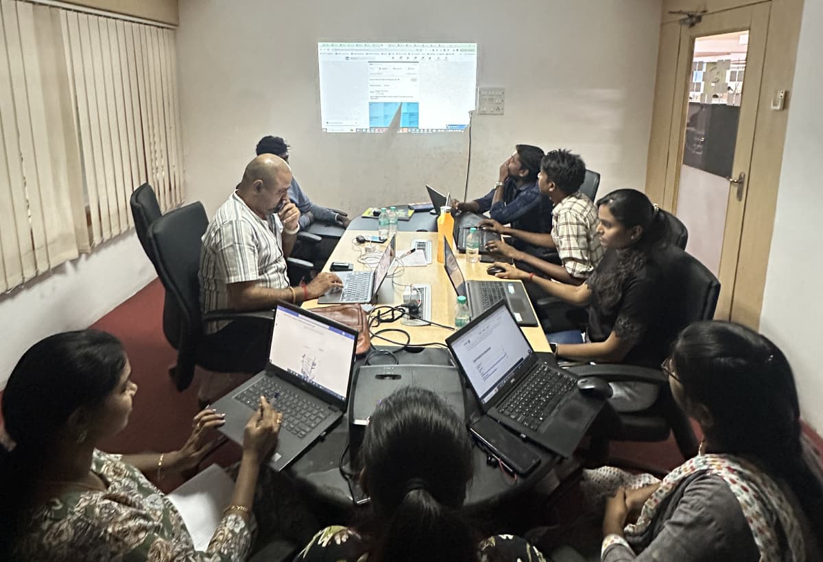 A working session around a boardroom table, eight people with laptops open and a screen projected on the wall